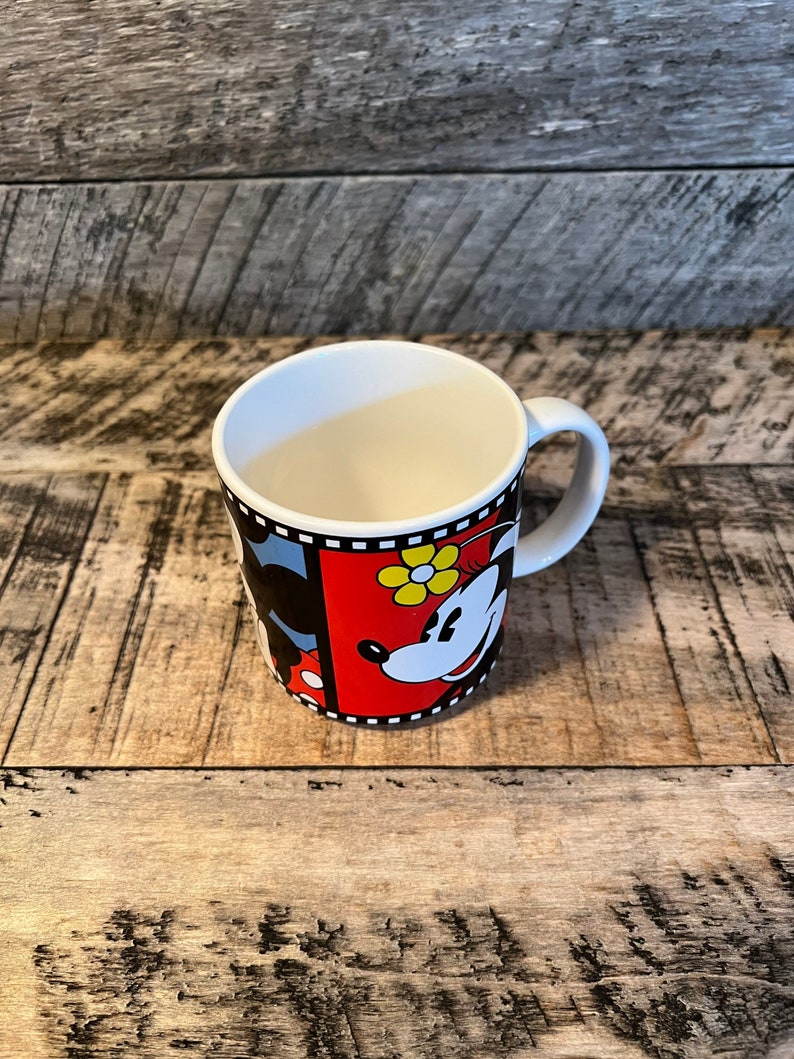 May include: A white ceramic mug with a black and red design featuring Mickey Mouse and Minnie Mouse. The mug has a black handle and a film strip border.