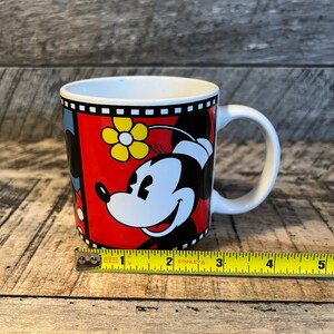 May include: A white ceramic mug with a red, black and yellow design featuring Mickey Mouse and Minnie Mouse. The mug has a black handle and a black rim.