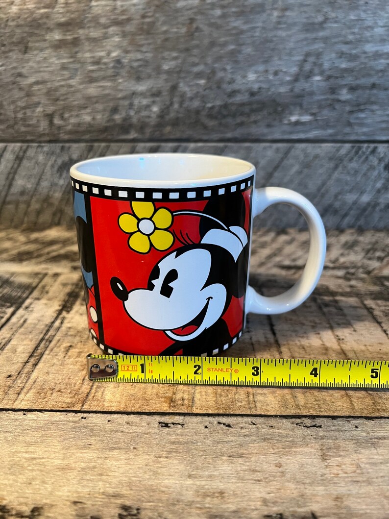 May include: A white ceramic mug with a red, black and yellow design featuring Mickey Mouse and Minnie Mouse. The mug has a black handle and a black rim.