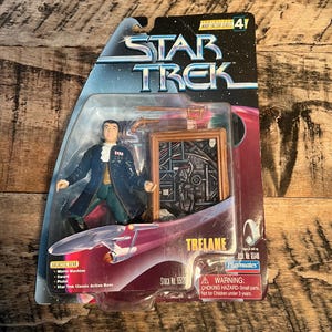May include: A Star Trek action figure of a man in a green and black outfit with a sword and pistol. The figure is packaged in a plastic blister pack with a red and silver background. The packaging includes the text "Star Trek" and "Trelene".