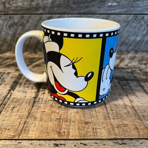 May include: White ceramic mug featuring a cartoon image of Mickey Mouse and Minnie Mouse in black, yellow, and blue. The image is framed with a black and white film strip.