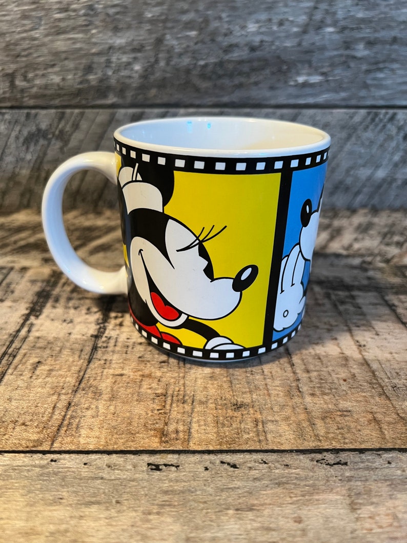 May include: White ceramic mug featuring a cartoon image of Mickey Mouse and Minnie Mouse in black, yellow, and blue. The image is framed with a black and white film strip.