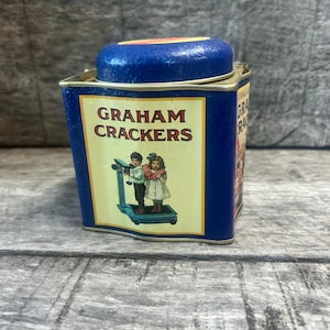May include: A vintage blue tin container for Graham Crackers. The tin features a rectangular label with the words "GRAHAM CRACKERS" and an illustration of two children. The lid is blue and the tin is rectangular.