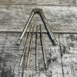 May include: Vintage silver nutcracker with four matching picks. The nutcracker is open, and the picks are arranged below it. The tools have a detailed, textured design and are made of a shiny, silver-colored metal. The background is a weathered wooden surface.