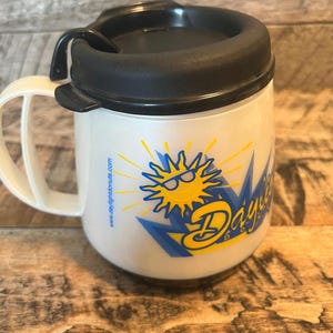 May include: White travel mug with a black lid and handle. The mug features a yellow and blue graphic of a sun wearing sunglasses, with the text "Daylight Donuts" in a stylized font. The website www.daylightdonuts.com is printed on the side.