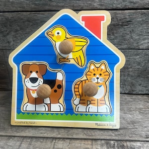 May include: A wooden house-shaped peg puzzle featuring a yellow bird, a brown and white dog, and an orange tabby cat. Each animal piece has a round wooden knob for grasping. The board includes the text ~crafted by hand~ and Melissa & Doug.
