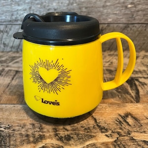 May include: A yellow travel mug with a black lid and handle. The mug features a heart design with radiating lines and the text "Love's". The mug is designed to keep beverages warm or cold.