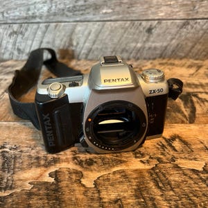 May include: A silver and black Pentax ZX-50 camera with a black strap. The camera has the Pentax logo on the front and side. The camera is on a wooden surface.