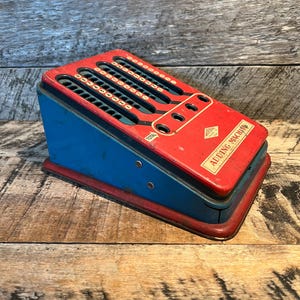 May include: Vintage adding machine in red, blue, and gold. The metal toy has a sloped design with rows of number wheels and the word "TOTAL" printed on the front. The label reads "ADDING MACHINE".