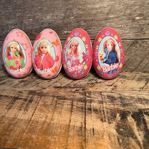 May include: Four pink Barbie-themed Easter egg containers. Each egg features a different Barbie image and the Barbie logo. The eggs are decorated with floral and butterfly patterns. The eggs are on a wooden surface.