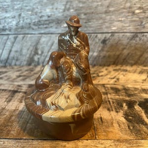 May include: A ceramic figurine depicting a man in a hat with two dogs. The figurine is brown and tan with a swirled pattern. The base is a round, lidded container. The background is a wooden surface.