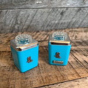 May include: Two blue salt and pepper shakers with silver tops and cowboy boot designs. The shaker on the right has the word "Astrodome" printed on it.