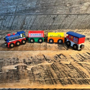 May include: A set of four colorful wooden toy train cars. The train cars are blue, red, yellow, and green. The first car is a blue engine with red wheels. The second car is red with the word "CARGO". The third car is yellow and orange. The last car is red and white.
