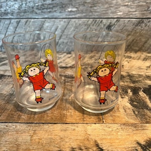 May include: Two clear drinking glasses with a colorful illustration of a girl jumping rope. The girl is wearing a red outfit and has blonde pigtails. The glasses are on a wooden surface.