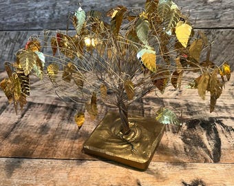 May include: A decorative metal tree sculpture with gold-colored leaves and a square base. The tree's trunk is a twisted wire, and the leaves have a metallic sheen. The base is painted gold, and the background is a wooden surface.