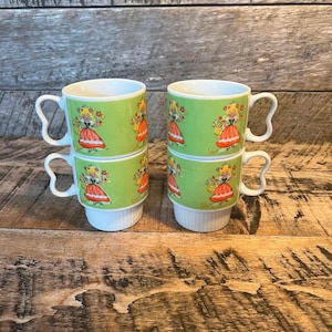 May include: Four stacked ceramic mugs with a green background and a design of a woman in a red dress carrying a basket of flowers. The mugs have white handles and bases, and are arranged on a wooden surface.