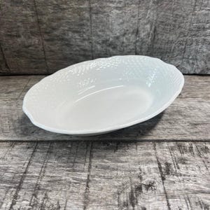 May include: A white, oval-shaped serving bowl with a textured rim. The bowl has a smooth, glossy interior and a slightly raised edge. The textured rim adds a decorative element to the otherwise simple design. The bowl is likely made of ceramic or porcelain.