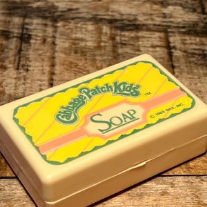 May include: A vintage Cabbage Patch Kids soap dish. The rectangular, off-white plastic container features a yellow and green label with the words "Cabbage Patch Kids" and "SOAP" in a retro font. The label has a pink border and the copyright date of 1983.