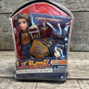 May include: A Lil' Boyz Koby doll in a clear plastic package. The doll has brown hair and blue eyes, wearing a yellow and black shirt and blue jeans. Accessories include a backpack and a red jacket. Text on the package reads "Real Mix 'N' Match Fashions Slip On 'N' Off!"