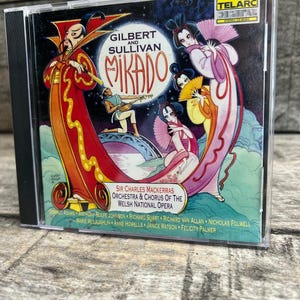 May include: A CD case featuring the Gilbert and Sullivan The Mikado album cover. The artwork displays stylized figures in traditional attire, including a man in red robes and women with fans, set against a moonlit background with text.