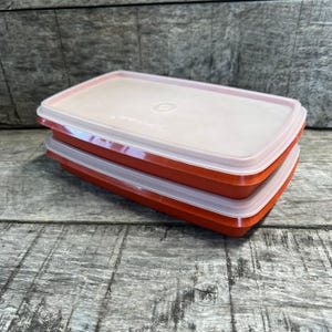 May include: Two rectangular storage containers stacked on top of each other. Each container features a solid orange base and a translucent, semi-clear lid. The lids have a circular indentation in the center and the word Tupperware embossed on them.