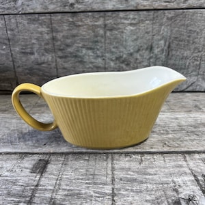 May include: A mustard yellow gravy boat featuring a ribbed exterior texture and a smooth cream-colored interior. The vessel includes a curved handle on one side and a wide pouring spout, showcasing a consistent color finish throughout the exterior.