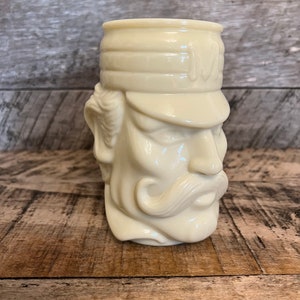 May include: A white ceramic mug shaped like a man's head wearing a hat. The mug has a mustache and a serious expression.