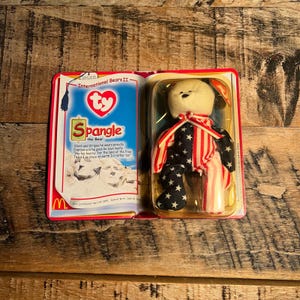 May include: A Ty Beanie Baby bear named Spangle, packaged in a clear plastic case. The bear is white with a red, white, and blue outfit. The packaging includes the text "International Bears II" and a poem about the bear.