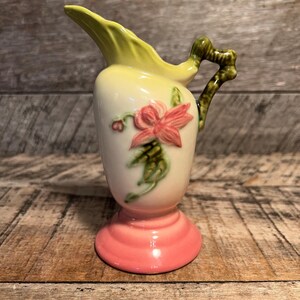 May include: A small, pink and yellow ceramic pitcher with a green handle and a floral design. The pitcher has a pink base and a yellow body with a green floral design.