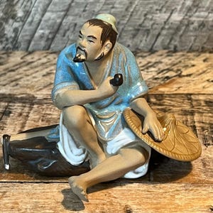 May include: A ceramic figurine of an Asian man in a blue robe, holding a pipe and a straw hat. The figurine is seated, with one leg bent and the other extended. The figure has a black beard and mustache.