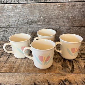 May include: Set of four white ceramic mugs with a floral design. The mugs feature pink, blue, and yellow stylized flowers.