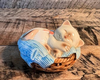 May include: A ceramic figurine of a sleeping cat. The cat is white with tan markings, resting on a blue blanket inside a brown woven basket. The cat has its eyes closed and a small red bow. The figurine is a decorative item.