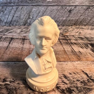 May include: A white ceramic bust of Mozart on a white base. The bust is detailed and shows Mozart's face and hair. The base has the word "MOZART" inscribed on it.