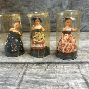 May include: Three miniature dolls in clear plastic tubes. Each doll is dressed in traditional folk attire, including colorful skirts and headwear. The dolls are standing on a dark base, and the background is a weathered gray surface.