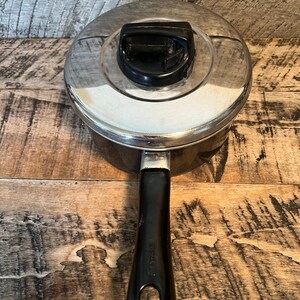 May include: A stainless steel saucepan with a lid and a black handle. The lid has a black handle on top. The handle is black and has a hole at the end for hanging. The pan is sitting on a wooden surface.