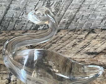 May include: A clear glass swan figurine. The swan has a long neck and a curved body. The figurine is sitting on a wooden surface.
