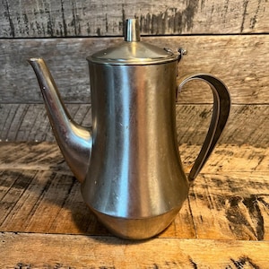 May include: A vintage silver-toned metal coffee pot with a curved spout, a hinged lid, and a curved handle. The pot has a bulbous body and sits on a wooden surface. The pot is likely used for serving coffee or tea.