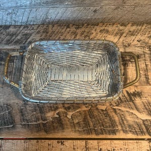 May include: Rectangular woven basket with silver-colored metal wire and brass-colored handles. The basket is empty and sits on a weathered wooden surface. The basket's design features a woven pattern throughout the body.