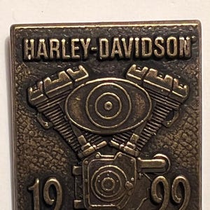 May include: A rectangular, bronze-colored Harley-Davidson emblem. The raised text "HARLEY-DAVIDSON" is at the top. Below, a detailed engine design is featured, with the year "1999" at the bottom. The emblem has a weathered, vintage appearance.