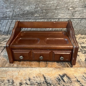 May include: A wooden jewelry organizer with a dark brown finish. It features three small drawers with ornate knobs and two recessed trays on top. The organizer has a curved back and is designed to sit on a dresser or table.