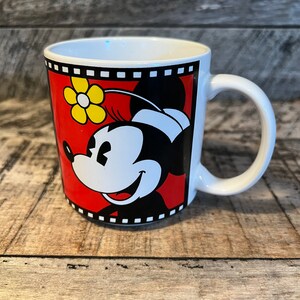 May include: White ceramic mug featuring a red, black and yellow image of Minnie Mouse. The image is framed with a black and white film strip design.