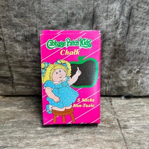 May include: A bright pink rectangular box of Cabbage Patch Kids chalk. The front features a cartoon character with blonde pigtails in a blue polka dot dress sitting on a stool, a black apple-shaped chalkboard, and the text 5 Sticks Non-Toxic.