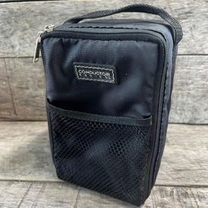 May include: A black rectangular storage bag with a top handle and a zipper closure. The front features a black mesh pocket and a rectangular patch with the white text CONDUCTOR SERIES. The bag is constructed from a durable, woven fabric material.
