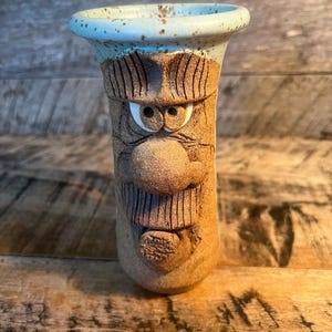 May include: A cylindrical ceramic mug featuring a sculpted face with a prominent nose, textured mustache, and wide eyes. The upper rim is glazed in a speckled light blue, while the lower portion has a natural, earthy brown clay finish.