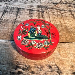 May include: A round, red decorative box with a lid featuring a painted scene of figures in traditional attire. The box is adorned with floral and tree motifs in various colors. The box is sitting on a wooden surface.