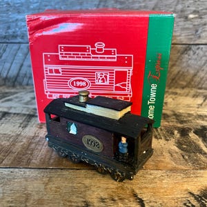 May include: A miniature train ornament with a maroon body and black roof, featuring the year "1998." A small figure stands inside. The ornament is in front of a red box with a train illustration and the words "Home Towne Express."