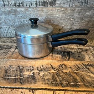 May include: A stainless steel double boiler with a black handle and lid knob. The pot is sitting on a wooden surface. The double boiler is designed for gentle cooking methods.