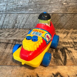 May include: A red and yellow toy race car with a blue driver figure. The car has a black helmet, blue wheels, and the words "Jumpin' Jimmy Racer" on the side. The car has a number "1" on the side.