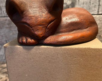 May include: A carved wooden cat figurine in a resting pose. The cat is a warm brown color with visible wood grain. The figurine is resting on a cardboard box, with the cat's paws tucked beneath its body.