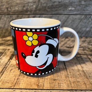 May include: A white ceramic mug with a black and red design featuring Mickey Mouse and Minnie Mouse. The design includes a yellow flower and a film strip border.